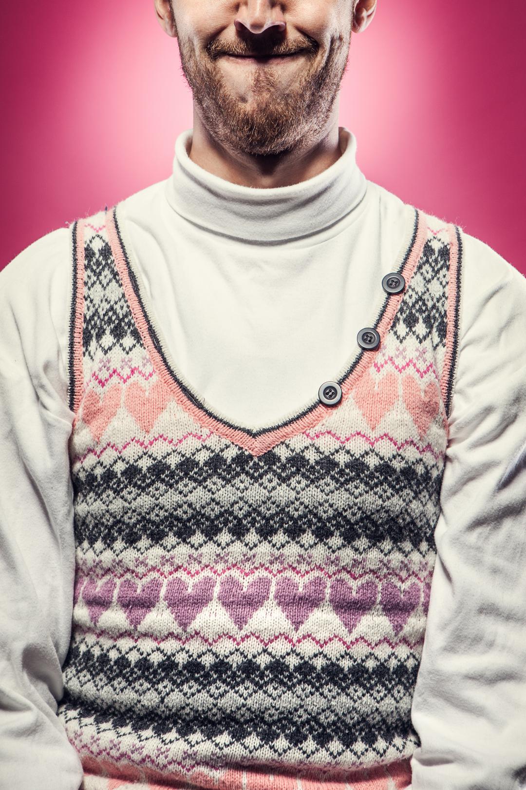 A man in a white turtleneck wears a wool sweater for Valentines day covered with knitted hearts. Vibrant pink background; studio shot.